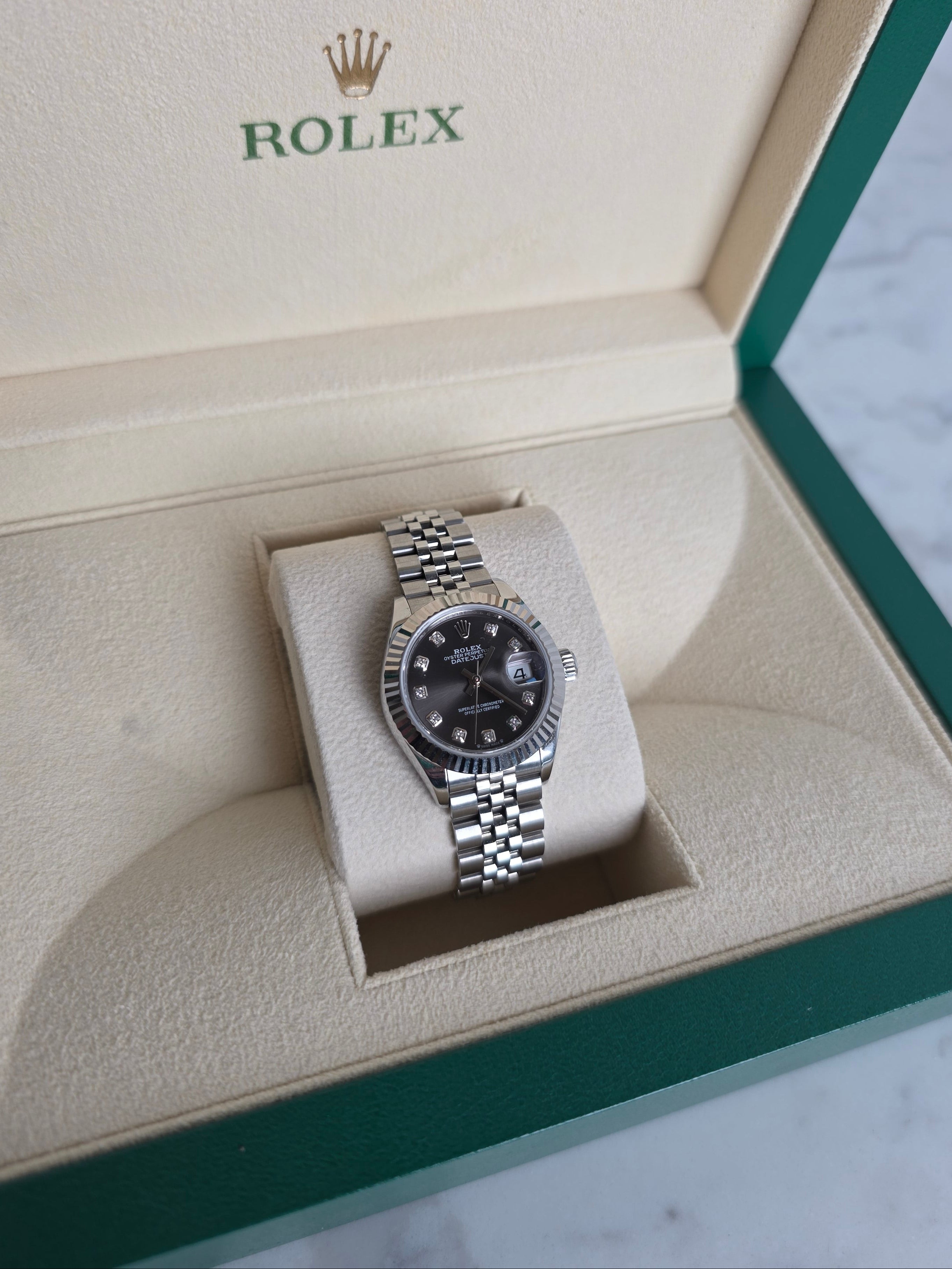 A Rolex wristwatch with a silver metal bracelet and black dial, displayed inside its open green and beige Rolex presentation box.
